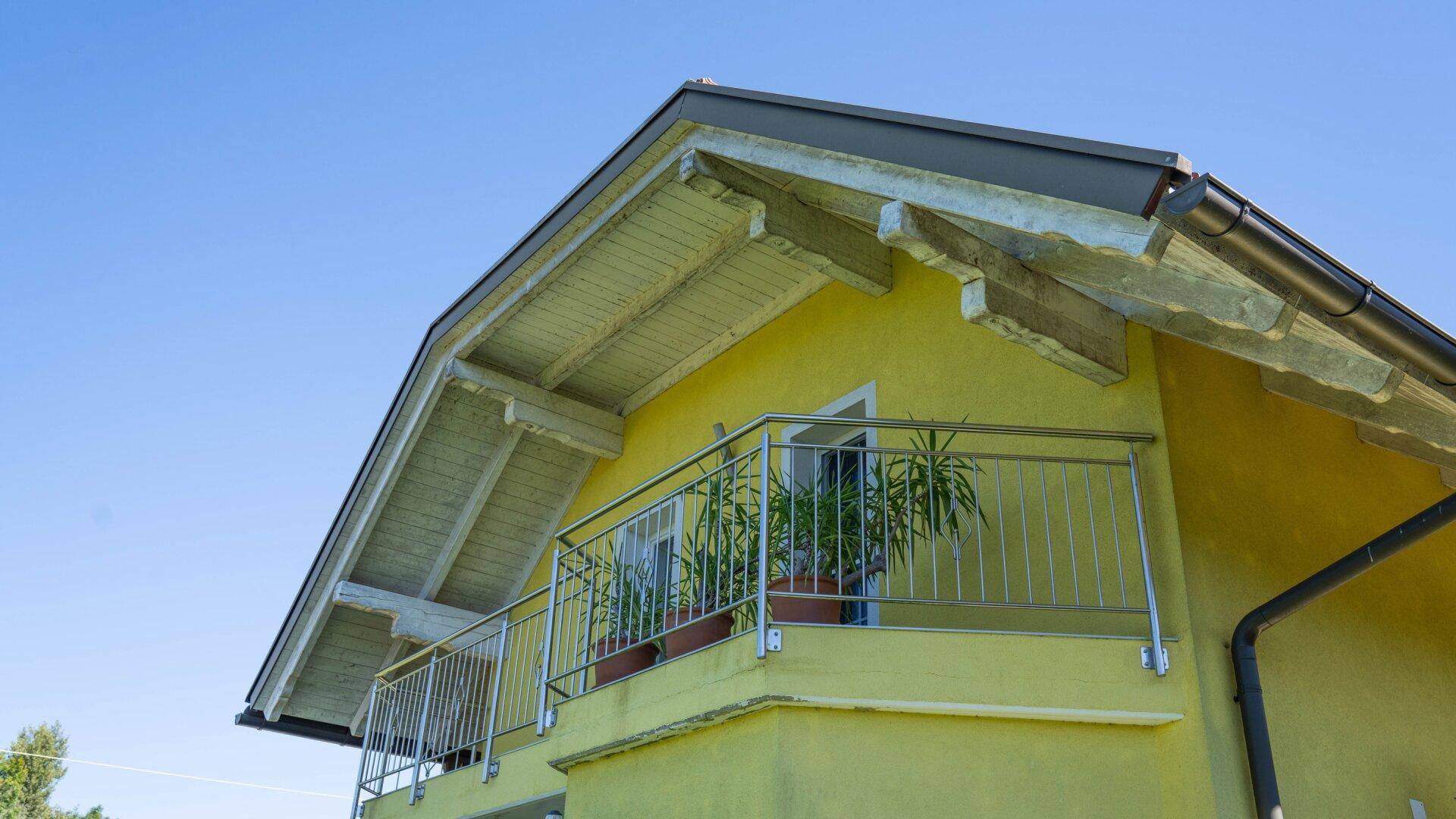 Gelbes Haus mit Giebeldach, Holzbalken und Balkon mit Metallgeländer.