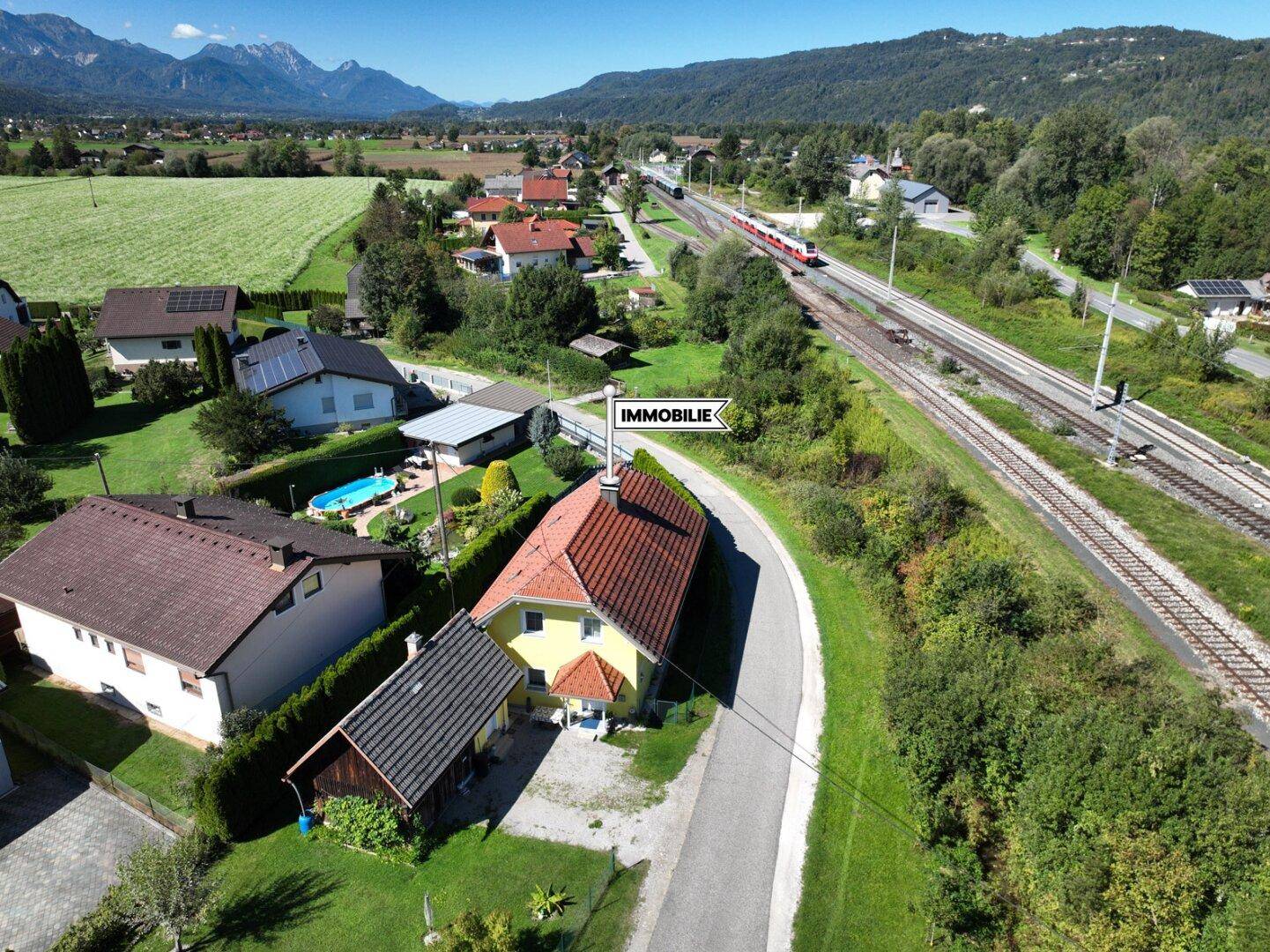 Luftaufnahme der Immobilie mit umliegenden Häusern, Feldern und Bahngleisen, umgeben von Bergen.