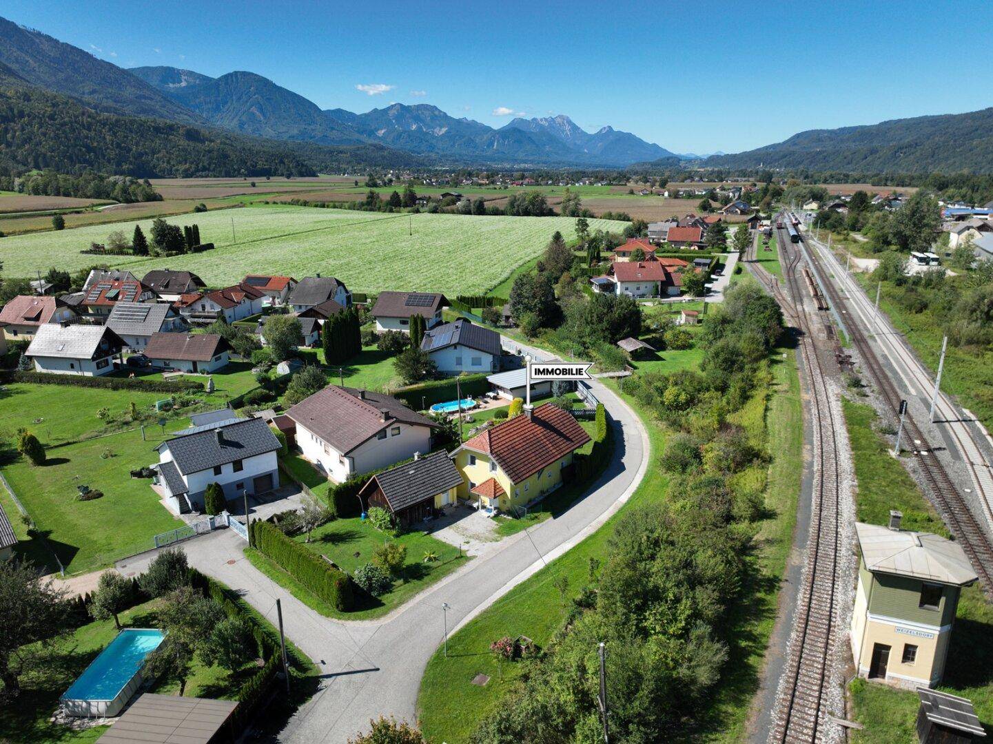 Luftaufnahme einer Wohnsiedlung mit Bergen im Hintergrund und Bahngleisen in der Nähe.