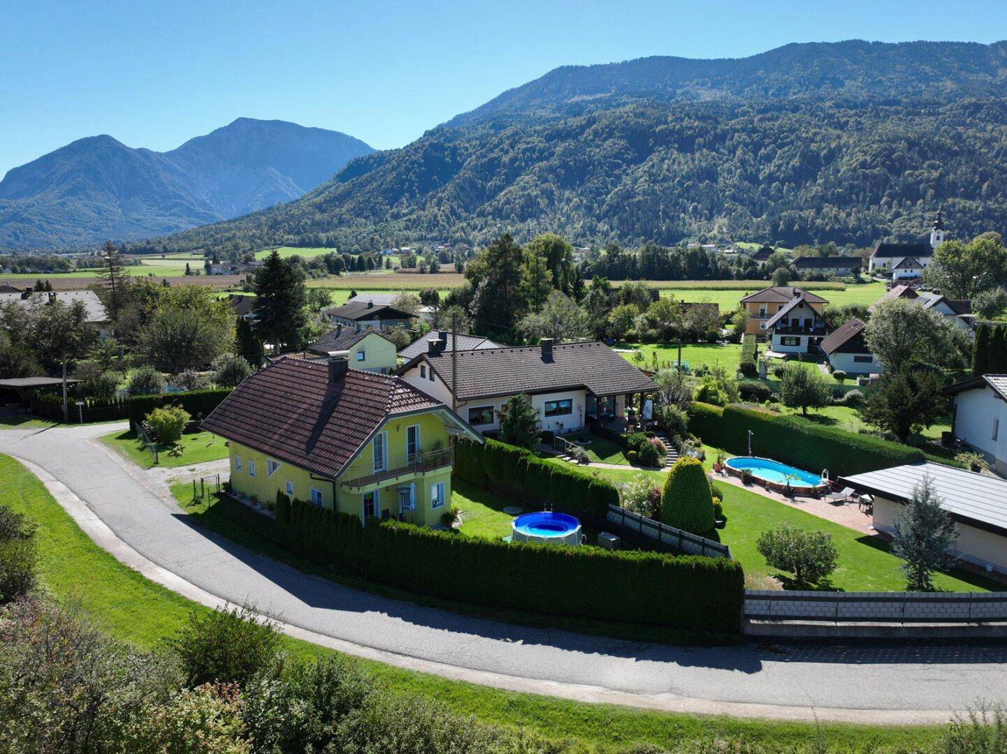 Luftaufnahme der Immobilie mit Garten, kleinem Pool und einem größeren Pool im Nachbargarten, umgeben von grüner Landschaft.