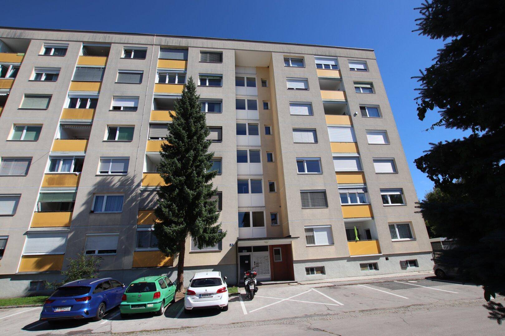 Mehrfamilienhaus mit mehreren Etagen, Balkonen und Parkplätzen im Freien.