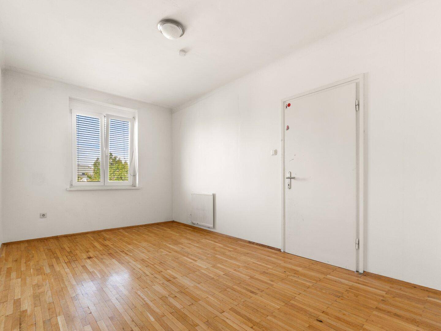 Helles Zimmer mit Parkettboden, weißen Wänden und einem Fenster mit Jalousien.