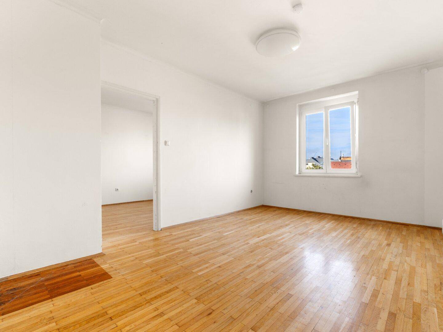 Freundliches Zimmer mit Parkettboden, weißen Wänden und Zugang zu einem weiteren Raum.