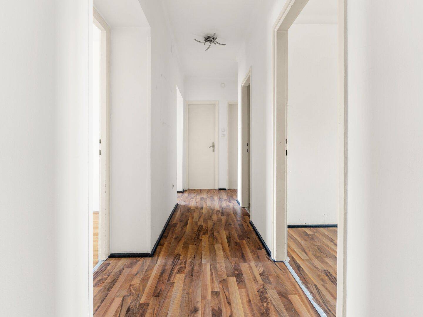 Langer Flur mit Laminatboden und weißen Wänden, der zu mehreren Zimmern führt.