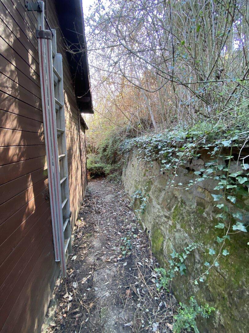 Schmaler Pfad entlang einer Holzwand und einer bewachsenen Steinmauer im Freien, umgeben von Natur.