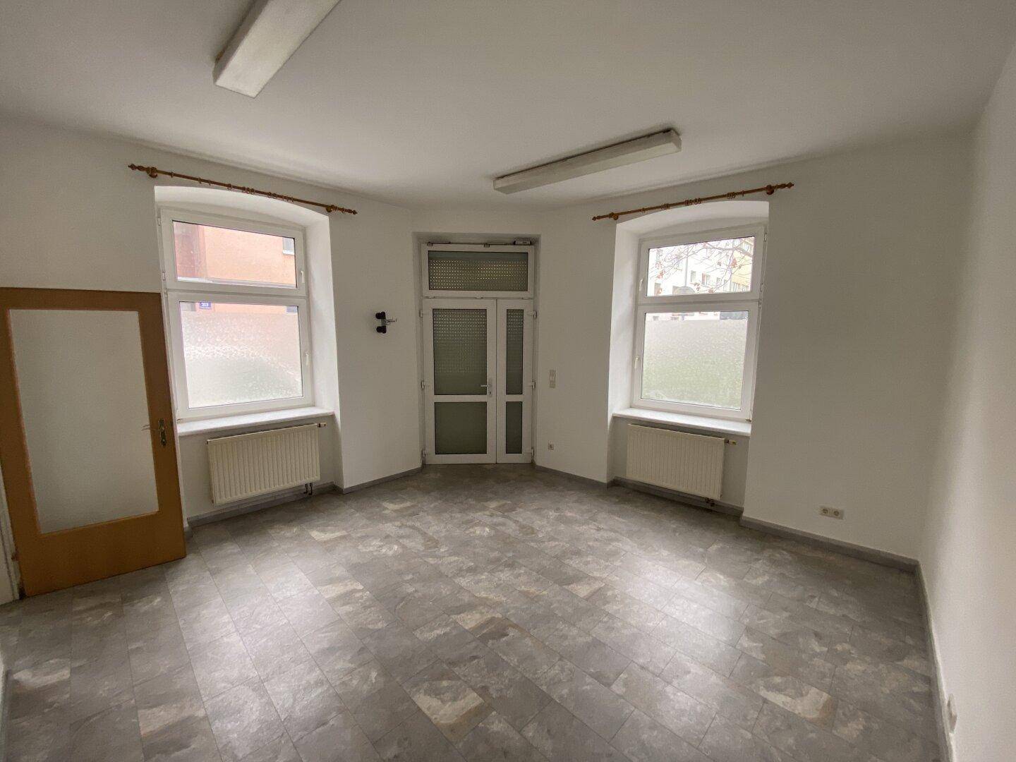 Heller Büroraum mit Eingangstür aus Glas, zwei Fenstern und grauem Vinylboden.