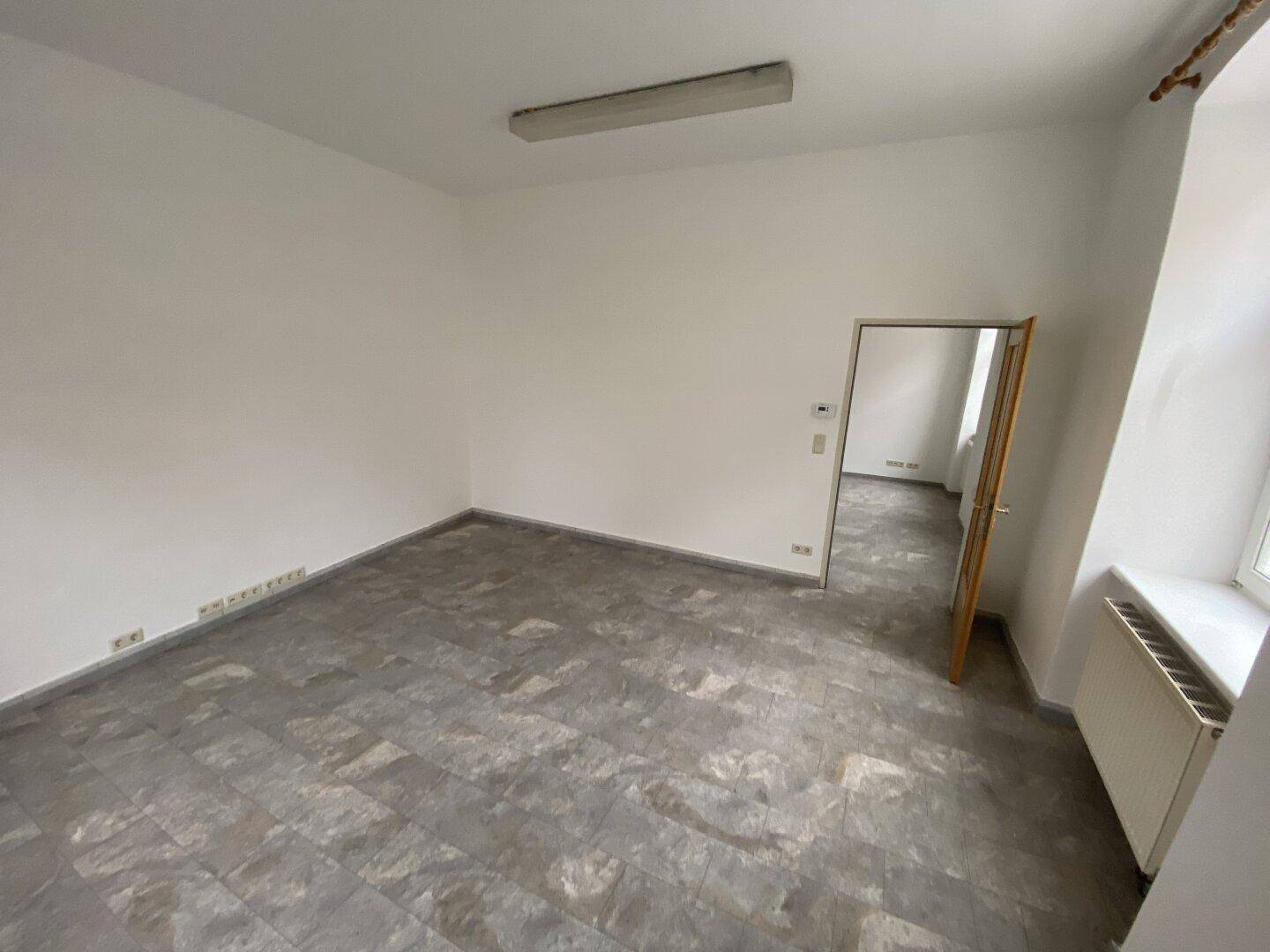 Leerer Büroraum mit grauem Vinylboden, weißen Wänden und einer Holztür.