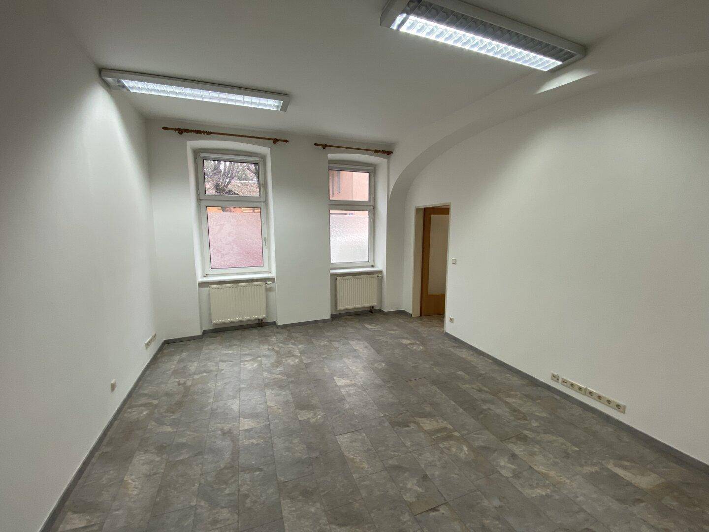 Heller Büroraum mit zwei Fenstern, weißen Wänden und grauem Vinylboden.