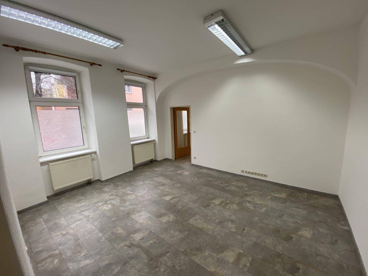 Heller Büroraum mit gewölbter Wand, zwei Fenstern und grauem Vinylboden.