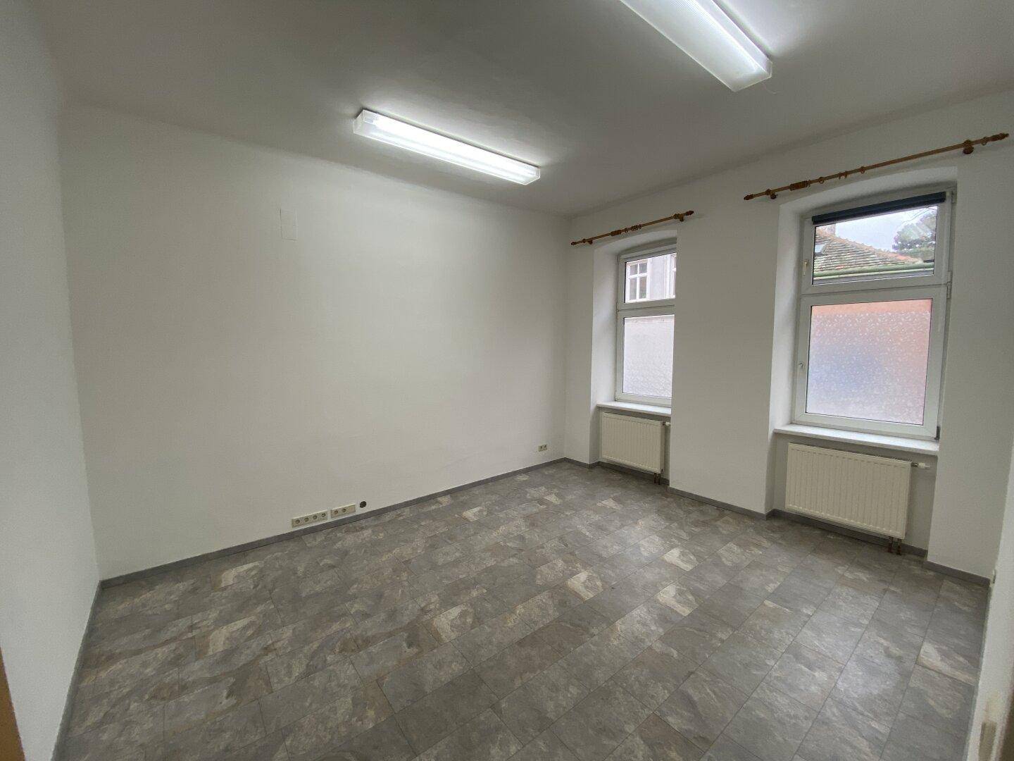 Geräumiger Büroraum mit zwei Fenstern, weißen Wänden und grauem Vinylboden.