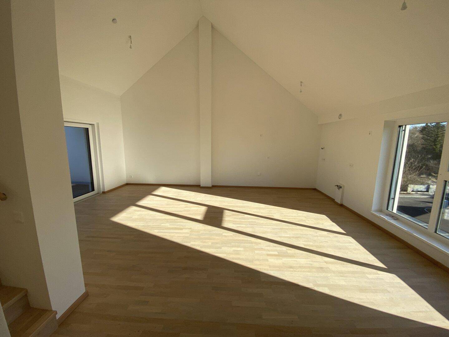 Geräumiger Wohnbereich mit schrägen Decken und großen Fenstern, die viel Sonnenlicht hereinlassen.