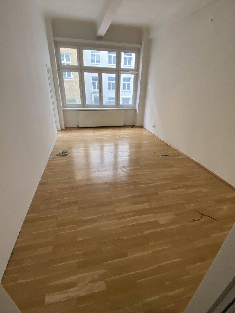 Heller, leerer Raum mit Holzparkett und großem Fenster, bietet viel Tageslicht und eine angenehme Atmosphäre.