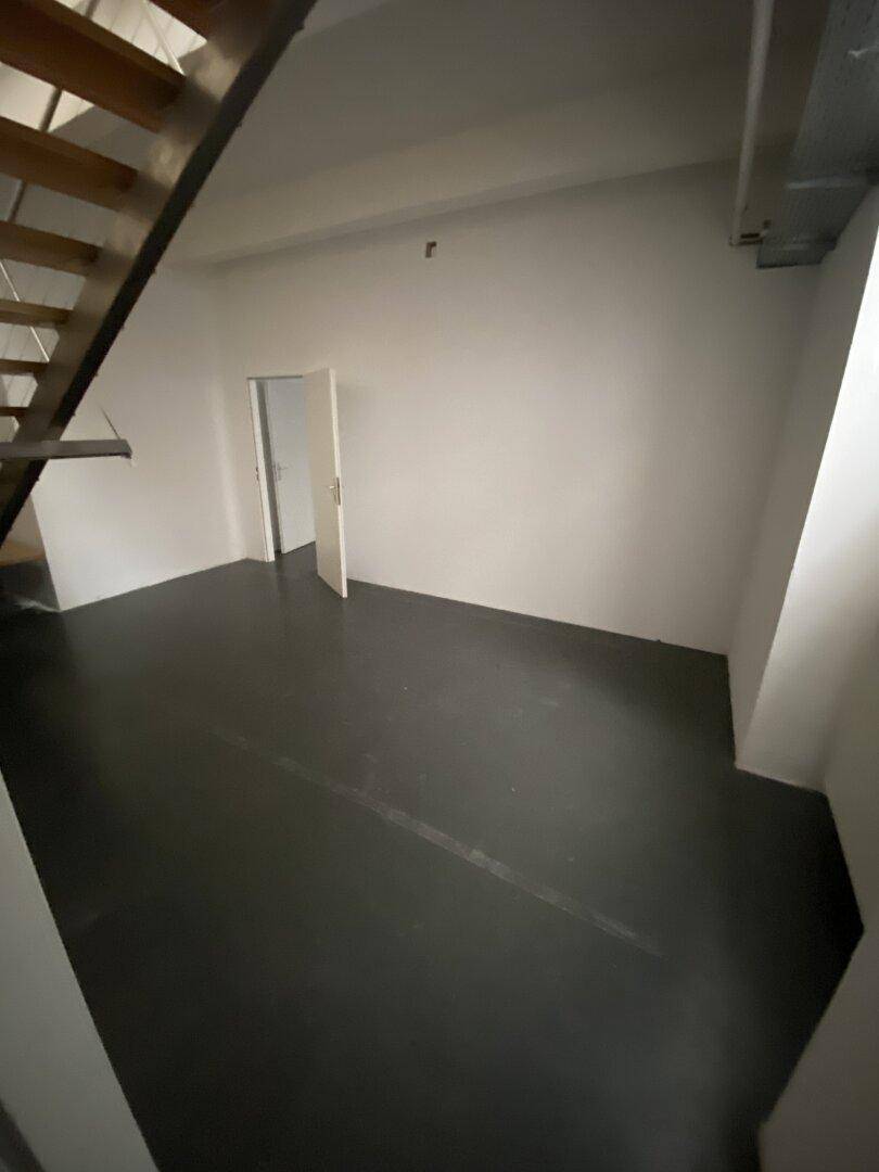 Raum mit dunklem Boden und offener Tür, daneben eine Treppe, die zu einer anderen Ebene führt.