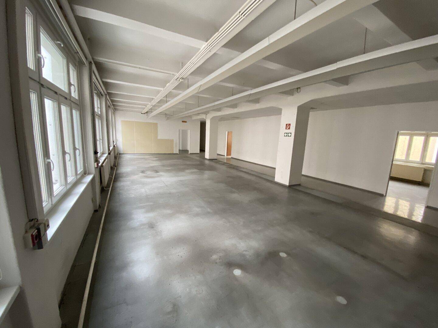 Geräumiger Bürobereich mit großen Fenstern, Betonboden und sichtbaren Deckenbalken, bereit zur individuellen Gestaltung.