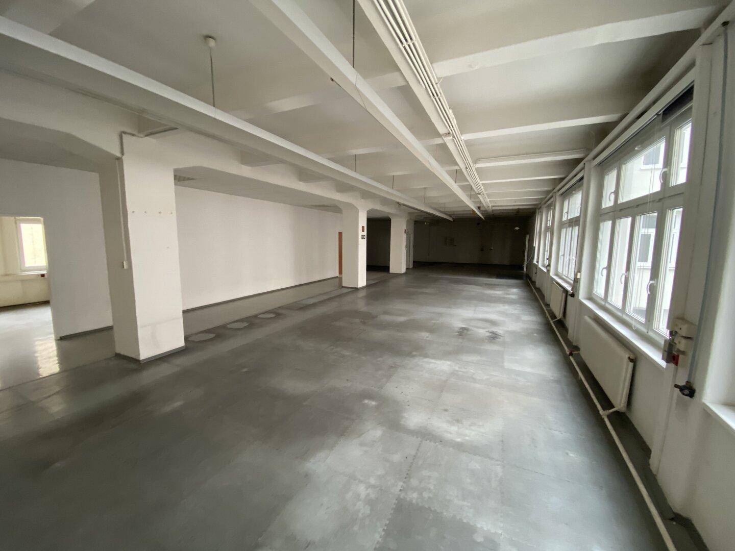 Weitläufiger Büroraum mit vielen Fenstern, Betonboden und Heizkörpern unter den Fenstern, bietet viel Tageslicht.