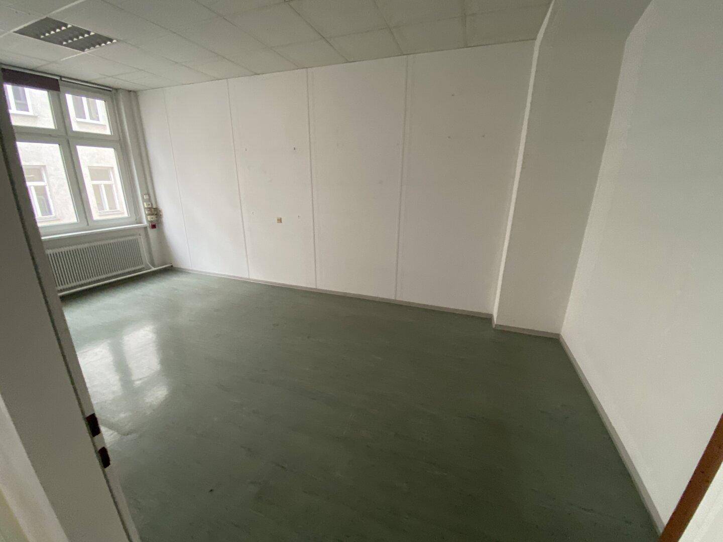 Kleinerer Büroraum mit Fenster, abgehängter Decke und grünlichem Bodenbelag, ideal für ein Einzelbüro.