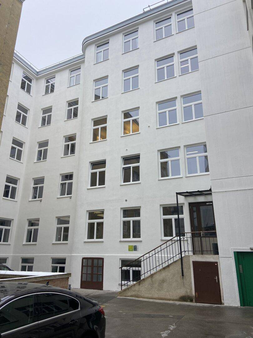 Weiße Gebäudefassade mit vielen Fenstern und einem Eingangsbereich mit Treppe, zeigt die Außenansicht.