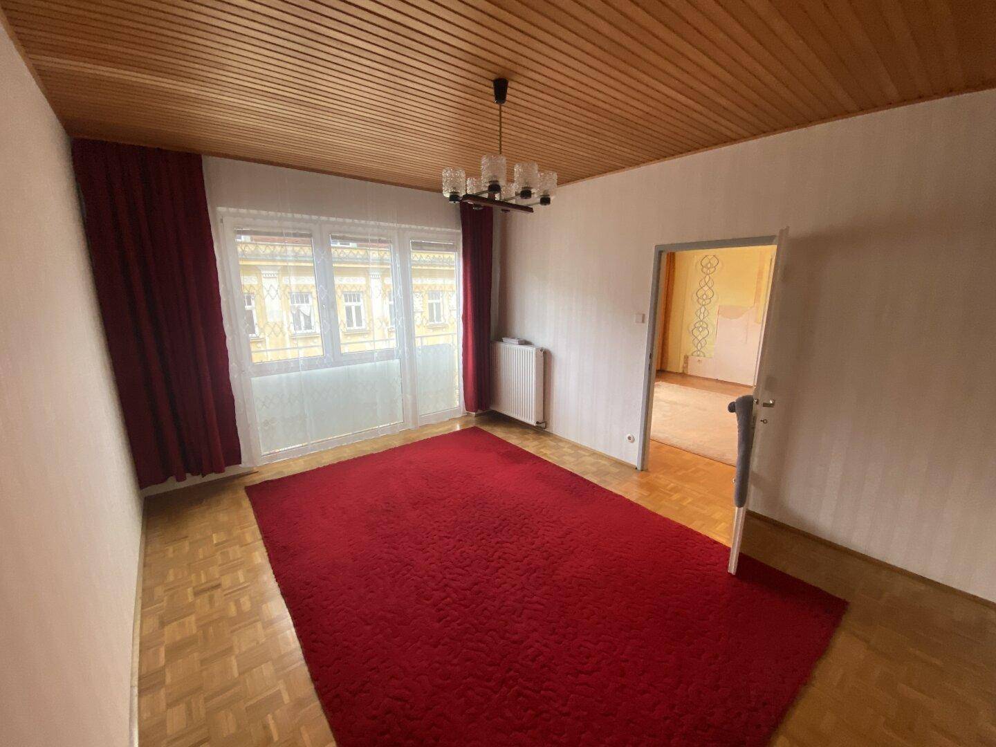 Geräumiges Wohnzimmer mit Parkettboden, Holzdecke und Zugang zu einem kleinen Balkon.