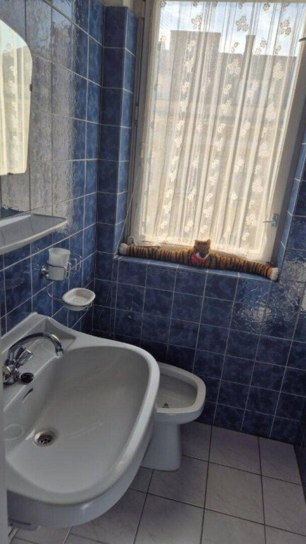 Badezimmer mit blauen Fliesen, Waschbecken, Bidet und Fenster mit Vorhang.