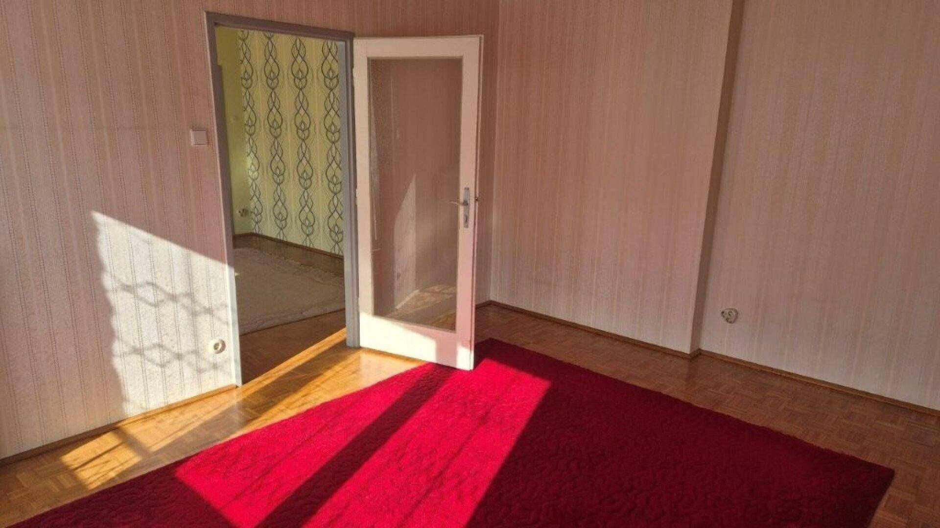 Leerer Raum mit Holzboden, rotem Teppich und offener Tür zu einem weiteren Zimmer mit alter Tapete.