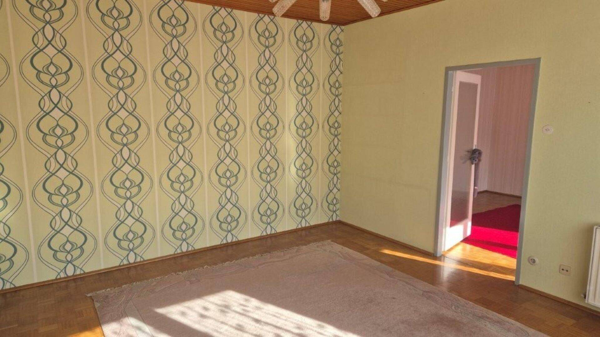 Wohnzimmer mit Parkettboden, Holzdecke und gemusterter Tapete an einer Wand.