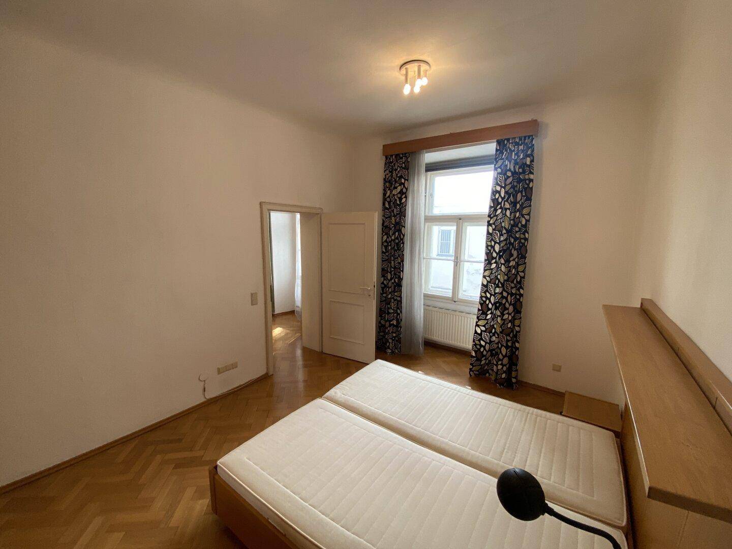 Schlafzimmer mit Doppelbett, Parkettboden und Fenster mit dunklen Vorhängen, das viel Licht hereinlässt.
