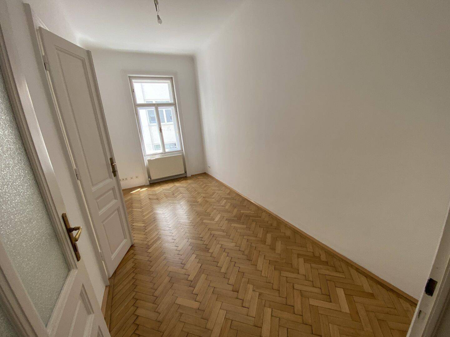 Helles Schlafzimmer mit Fischgrätparkett und einem großen Fenster, das viel Licht spendet.