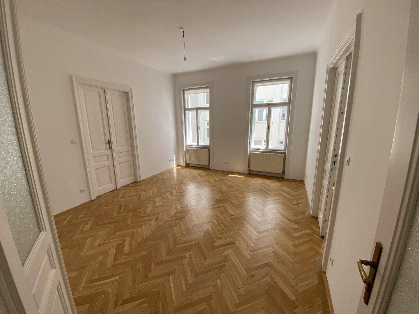 Geräumiges Wohnzimmer mit Fischgrätparkett und zwei großen Fenstern, die den Raum erhellen.