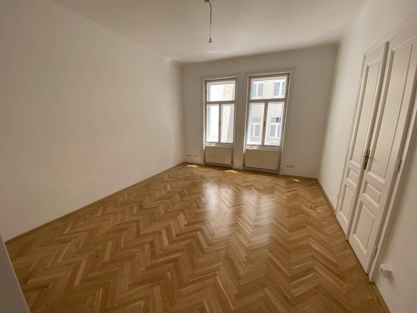 Helles Schlafzimmer mit Fischgrätparkett und zwei Fenstern, die den Raum mit Tageslicht füllen.