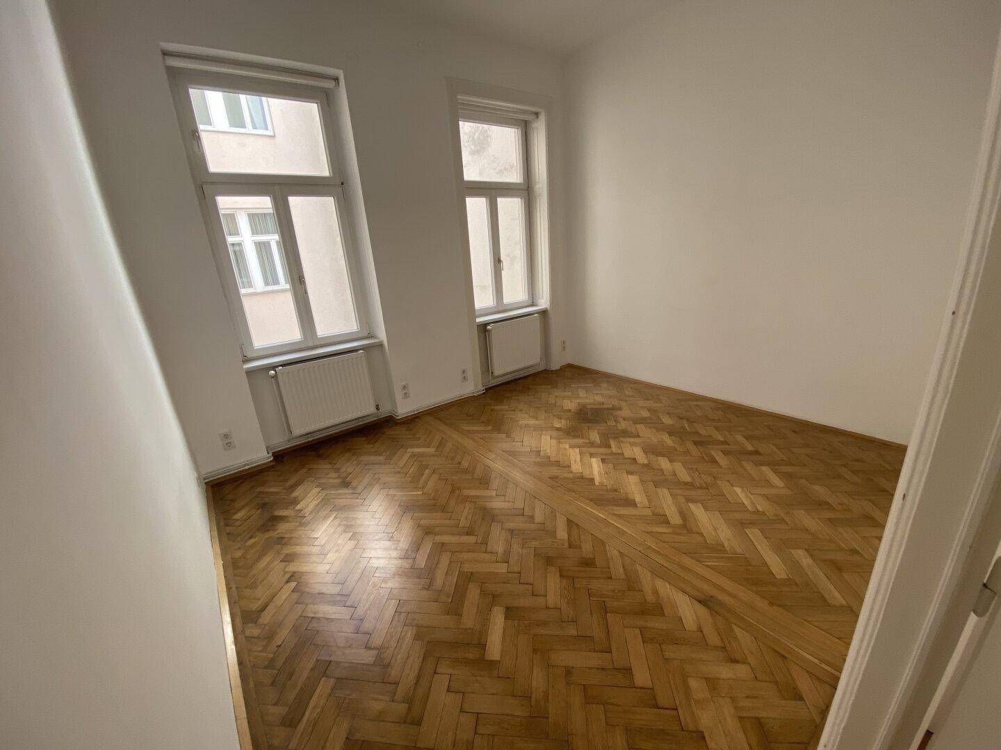 Helles Schlafzimmer mit Fischgrätparkett und zwei Fenstern, die den Raum mit Tageslicht durchfluten.