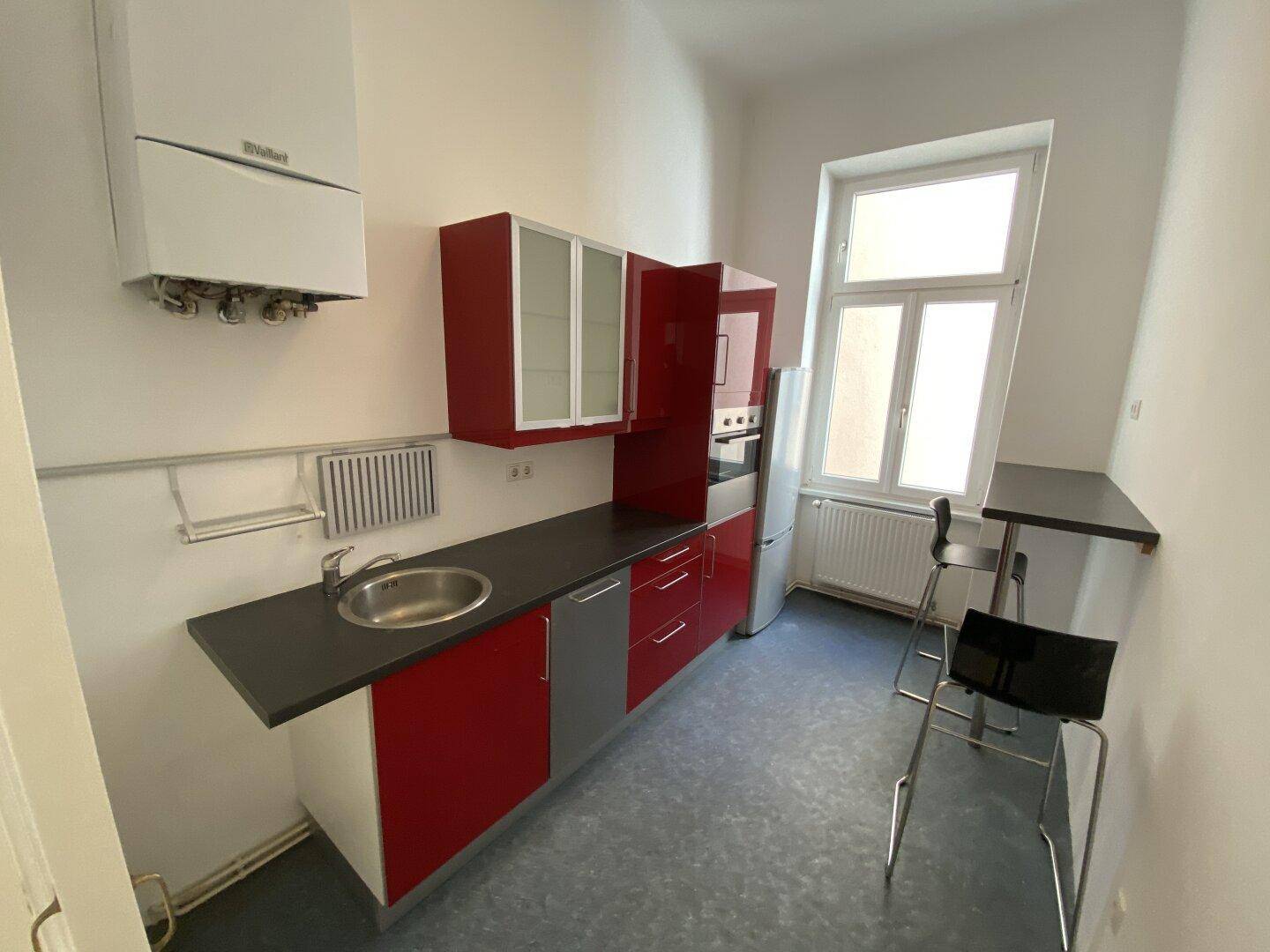 Küche mit roten Unterschränken, Spüle, Kühlschrank und einem Fenster, das Tageslicht hereinlässt.
