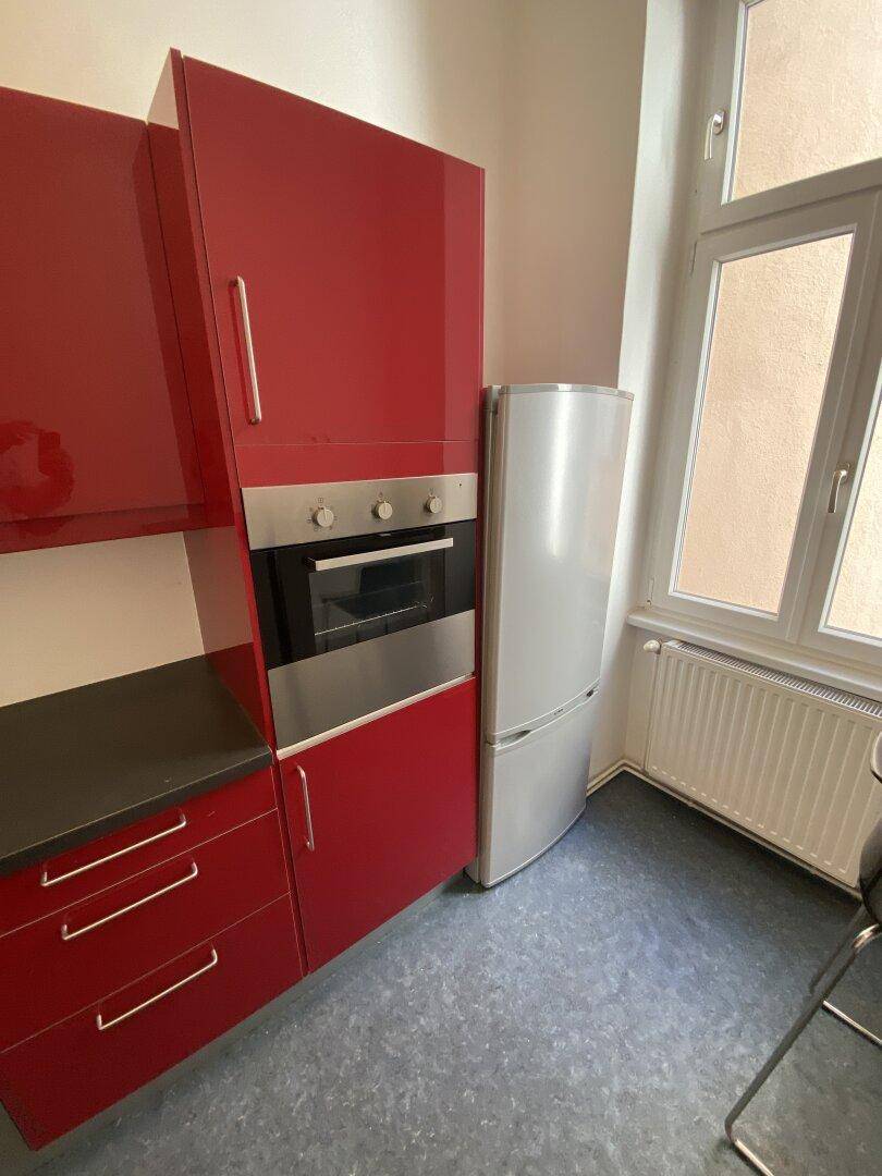 Einbauküche mit rotem Hochschrank, integriertem Backofen und freistehendem Kühlschrank neben dem Fenster.