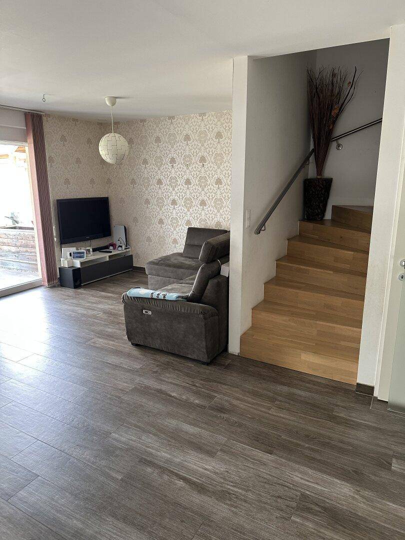 Geräumiges Wohnzimmer mit Ecksofa und Fernseher, mit offener Treppe zum Obergeschoss.