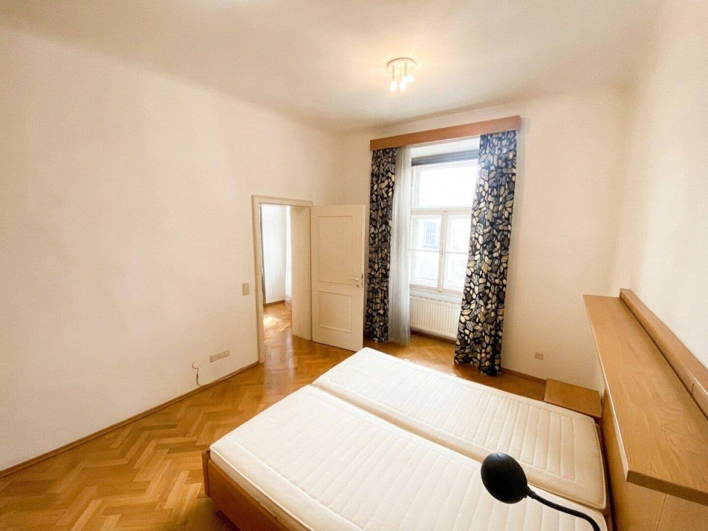 Gemütliches Schlafzimmer mit Doppelbett, Parkettboden und einem Fenster mit dunklen, gemusterten Vorhängen.