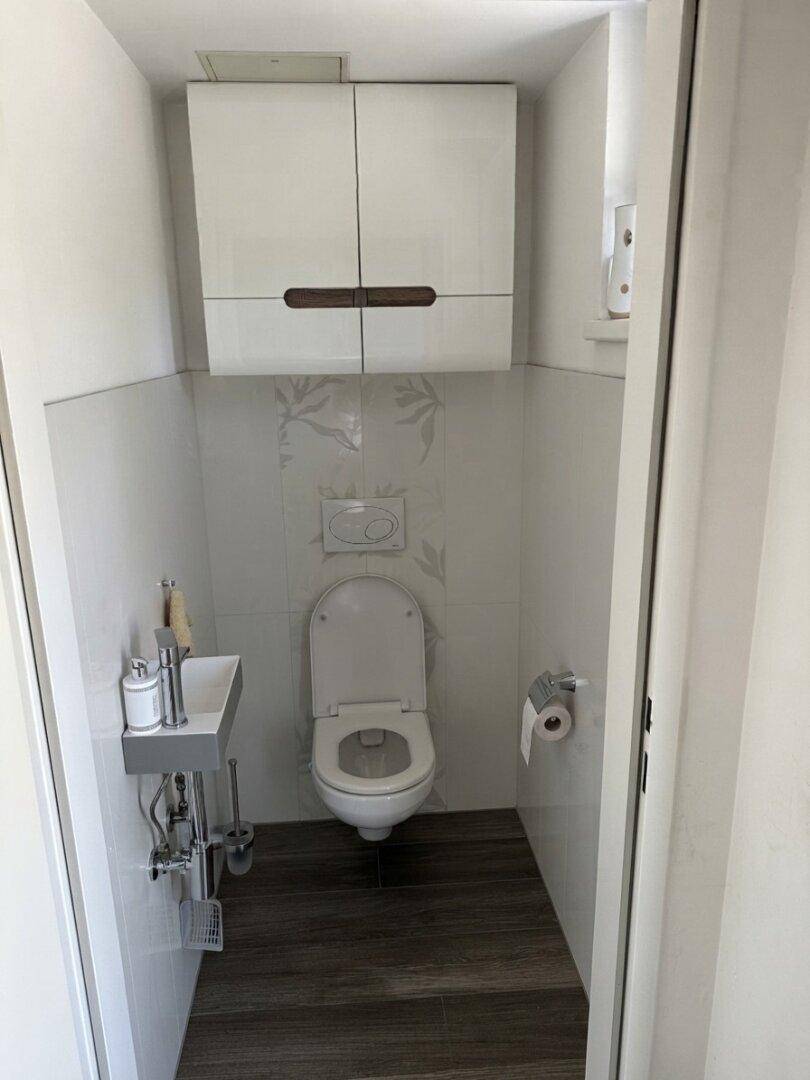 Kleines Gäste-WC mit Wand-WC, kleinem Waschbecken und weißen Hängeschränken.