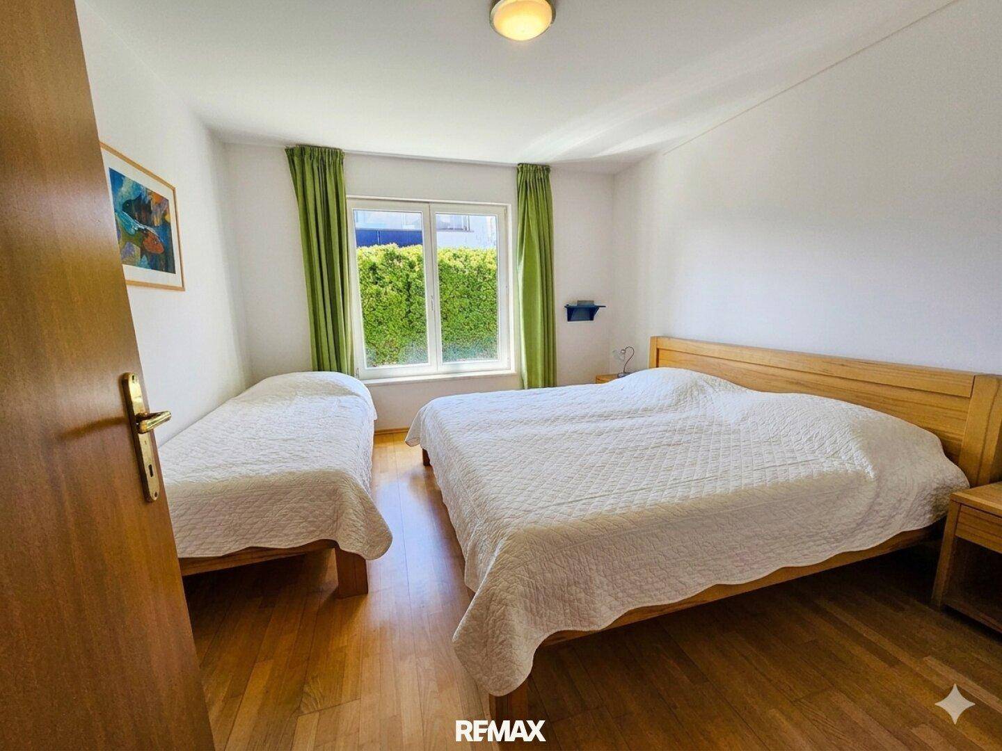 Schlafzimmer mit Doppelbett und Einzelbett, Holznachttisch und Blick auf eine Hecke.
