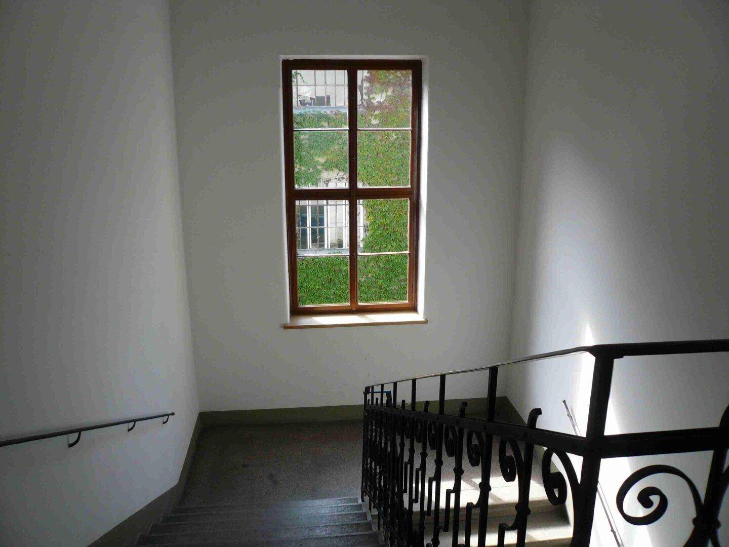 Treppenhaus mit schmiedeeisernem Geländer und einem Fenster mit Blick auf grüne Pflanzen im Außenbereich.