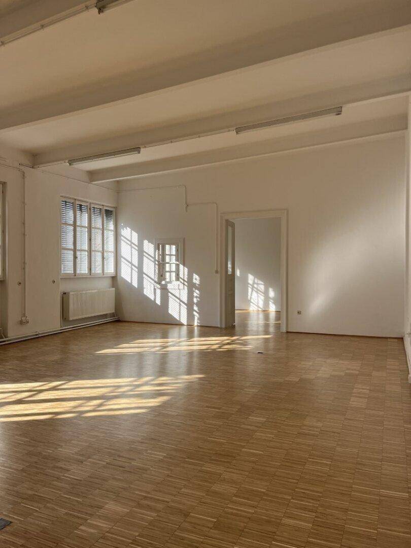 Großer, leerer Raum mit Holzboden, weißen Wänden und viel Sonnenlicht, das durch die Fenster fällt.