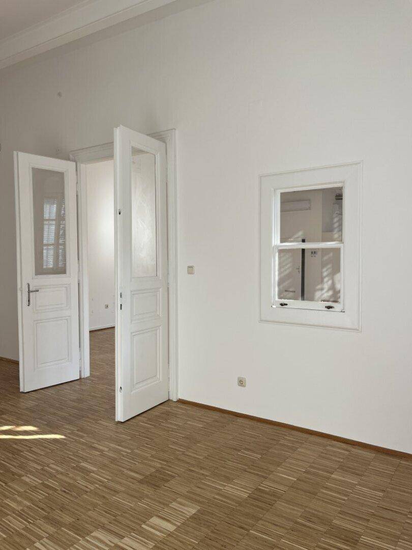 Leerer Raum mit Holzboden, weißen Wänden und Doppeltüren, die zu einem weiteren hellen Raum führen.