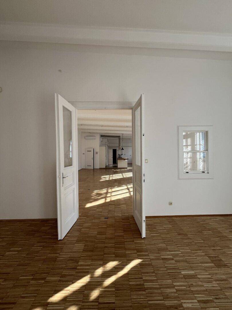 Geräumiger Wohnbereich mit Holzboden, hohen Decken und Doppeltüren, die zu weiteren Räumen führen.
