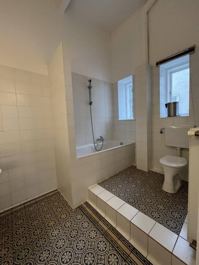 Helles Badezimmer mit Badewanne, Toilette und dekorativen Bodenfliesen, ausgestattet mit einem Fenster.