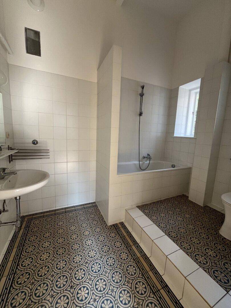 Badezimmer mit Badewanne, Waschbecken und gemusterten Fliesen auf dem Boden, inklusive Fenster und Abluftventilator.