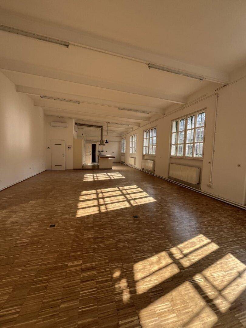 Heller, offener Wohnbereich mit Holzboden, hohen Decken und großen Fenstern, die viel Sonnenlicht hereinlassen.