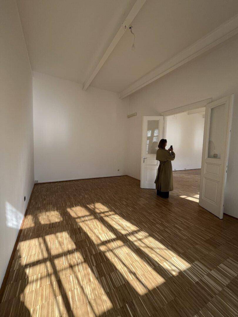 Heller, leerer Raum mit Holzboden und einer Person, die den Raum fotografiert, mit viel natürlichem Licht.