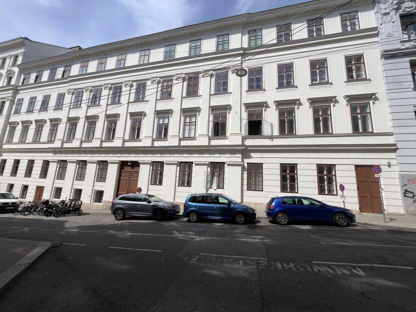 Klassische Fassade eines Mehrfamilienhauses mit vielen Fenstern und parkenden Autos auf der Straße.