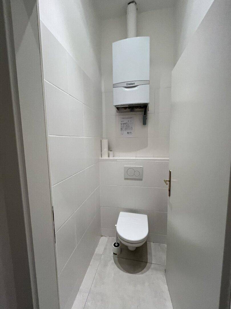 Gäste-WC mit wandhängender Toilette, weißen Fliesen und einem an der Wand montierten Warmwasserbereiter.