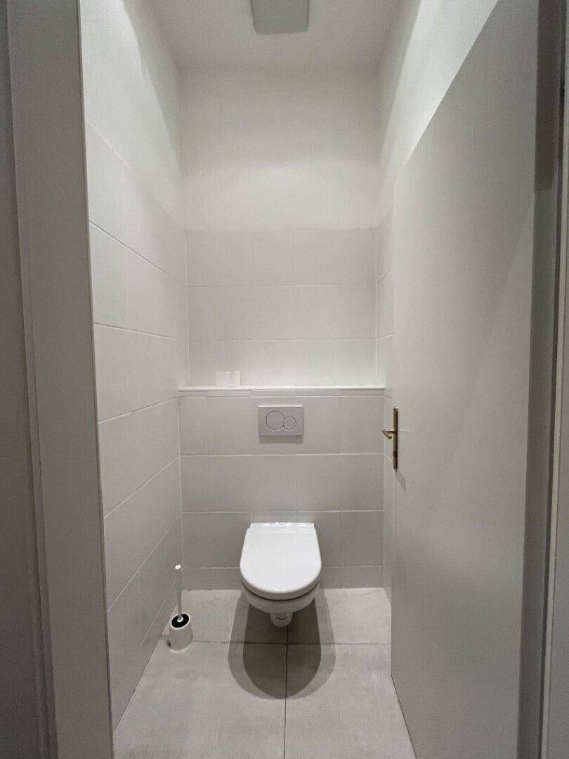Modernes Gäste-WC mit wandhängender Toilette und weißen Fliesen an den Wänden und auf dem Boden.