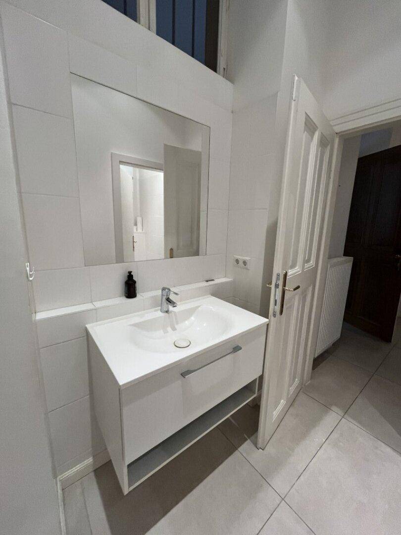 Modernes Badezimmer mit weißem Waschtisch, großem Spiegel und hellen Fliesen an Wänden und Boden.
