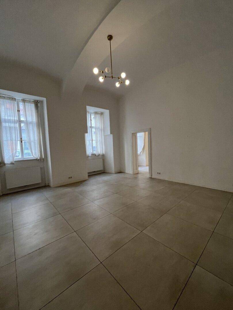 Geräumiger Raum mit hohen Decken, zwei Fenstern mit Vorhängen und einem modernen Fliesenboden.
