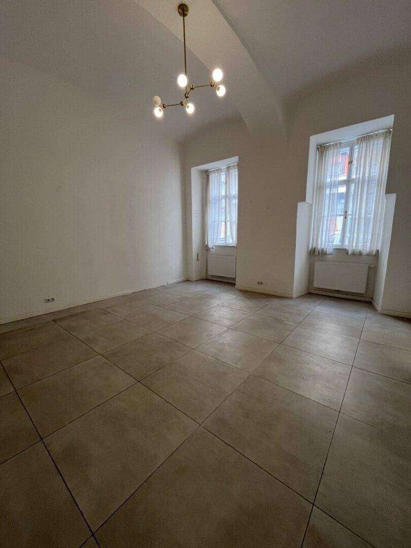 Heller Wohnraum mit hohen Decken, zwei Fenstern und einem modernen Fliesenboden in neutralen Farben.